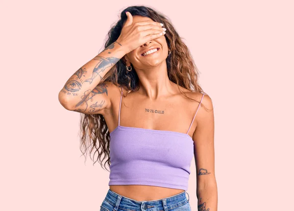Young hispanic woman with tattoo wearing casual clothes smiling and laughing with hand on face covering eyes for surprise. blind concept.
