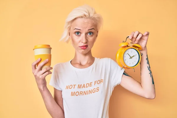 目覚まし時計を持っている入れ墨を持つ若いブロンドの女性とコーヒーのカップを取る無知で混乱した表情 疑わしい概念 — ストック写真