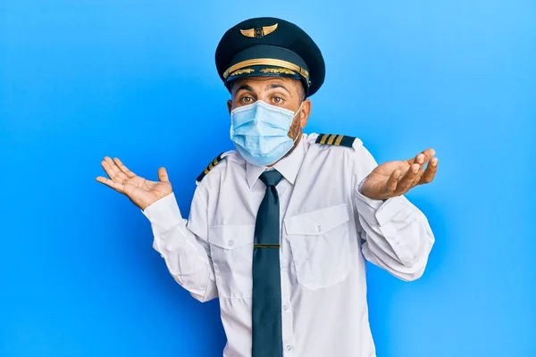 髭を生やしたハンサムな男は 安全マスクを身に着けている飛行機のパイロット制服を着て 腕や手を上げて混乱した表情 疑わしい概念 — ストック写真