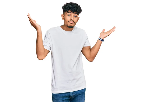 年轻的阿拉伯男子穿着随意的白色T恤 毫无头绪 满脸困惑 手臂和双手高举 怀疑概念 — 图库照片