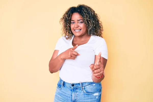 若いアフリカ系アメリカ人のプラスサイズの女性が幸せと面白い顔を持つカメラに指を指してカジュアルな白いTシャツを着て 良いエネルギーと振動は — ストック写真