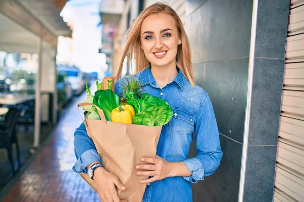年轻的金发姑娘高兴地微笑着 手里拿着食品杂货纸袋站在城里 — 图库照片