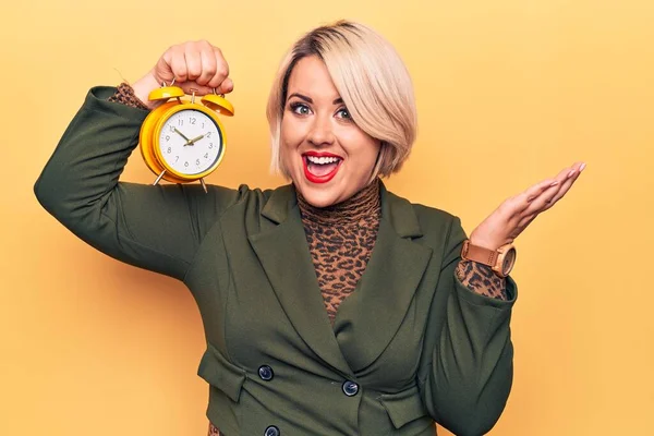 Young beautiful plus size blonde woman holding vintage alarm clock over yellow background celebrating achievement with happy smile and winner expression with raised hand