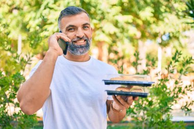 Orta yaşlı, kır saçlı, akıllı telefondan konuşan ve elinde yemek tutan bir adam.