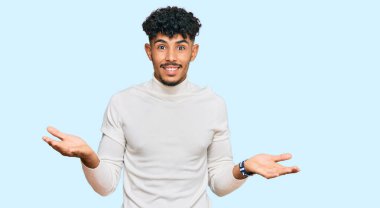 Genç Arap adam sıradan bir kazak giyiyor. Neşeli gülümsüyor ve kollarını açıyor. Hoş geldin, olumlu ve kendinden emin selamlar. 