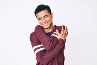 Beyaz arka planda genç, yakışıklı İspanyol bir adam kendine sarılıyor mutlu ve pozitif, kendinden emin gülümsüyor. Sevgi ve özgüven. 