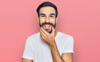 Gündelik beyaz tişört giyen genç İspanyol adam kameraya kendinden emin bakıyor kollarını kavuşturmuş ve çenesini kaldırmış gülümsüyor. Olumlu düşünmek. 