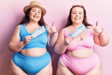 Bikini giyen, mutlu ve pozitif gülümseyen, başparmağıyla mükemmel ve onay işareti yapan genç ve büyük beden ikizler. 