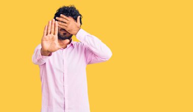 Kıvırcık saçlı yakışıklı genç adam ve ayı iş kıyafetleri giyip gözleri ellerle kapatıyor ve üzgün ve korku ifadeleriyle jest yapıyor. utangaç ve olumsuz kavram. 