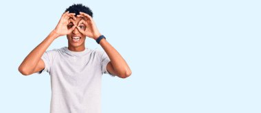 Genç Afro-Amerikan bir adam günlük kıyafetler giyiyor dürbün gibi jest yapıyor, dili dışarı çıkarıyor, gözleri parmaklarından bakıyor. çılgın ifade. 