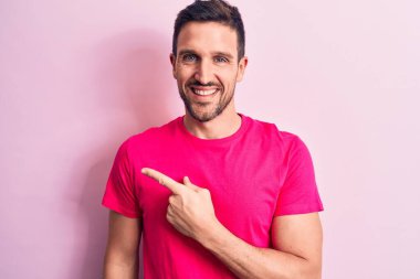 Genç ve yakışıklı bir adam, günlük tişört giyip pembe arka planda dikiliyor. Gülümsüyor, gülümsüyor, elini ve parmağını yana kaldırıyor.