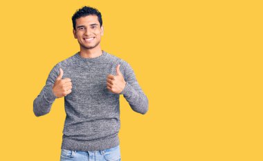 İspanyol asıllı yakışıklı genç adam, günlük süveter giyer, başarı işareti yapar, elleriyle olumlu jest yapar, gülümser ve mutlu olur. Neşeli ifade ve kazanan jest. 