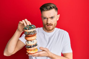 Genç kızıl saçlı adam elinde lezzetli renkli çörekler tutuyor. Hiçbir şeyden haberi yok ve kafası karışık. şüphe kavramı. 