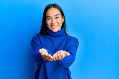 Asyalı genç bir kadın, sıradan bir kazak giyip avuçlarını birbirine değdirip gülümseyerek jest yapıyor. Bekleme ve koruma 