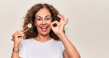 Orta yaşlı güzel bir kadın beyaz arka planda tahta çubuklar kullanarak suşi yiyor. Parmaklarıyla işaret veriyor, gülümseyerek mükemmel bir sembol oluşturuyor.