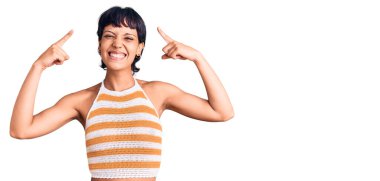 Kısa saçlı genç esmer kadın. Günlük kıyafetler giyiyor. İki eliyle başını gösteren gülümsüyor. 