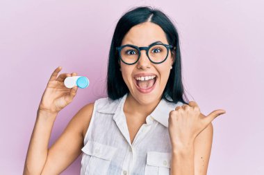 Güzel, genç bir kadın, elinde gözlük ve lens tutarken başparmağıyla ağzını açıp gülümsüyor. 