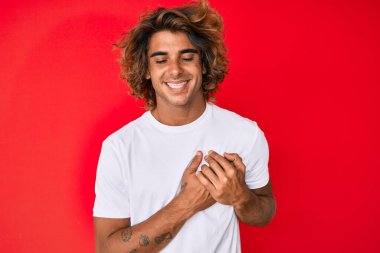 Gündelik beyaz tişört giyen, elleri göğsünde, gözleri kapalı gülümseyen ve yüzünde minnet ifadesi olan genç İspanyol bir adam. sağlık kavramı. 