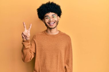 Afro saçlı, sıradan bir kazak giyen Afro Amerikalı genç adam iki numarayı gösterip gülümserken kendinden emin ve mutlu görünüyor.. 