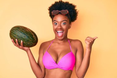 Genç Afro-Amerikan kadın bikini giyiyor elinde kavun başparmağıyla yan tarafa bakıyor mutlu bir şekilde gülümsüyor. 