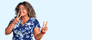 Genç Afro-Amerikan artı cüsseli kadın günlük kıyafetler giyiyor gülümsüyor kameraya bakıyor zafer işareti yaparken parmaklarını gösteriyor. İki numara.. 