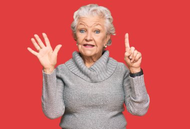 Kış süveteri giyen yaşlı, gri saçlı kadın altı numaralı parmaklarını gösterip gülümserken mutlu ve kendinden emin.. 
