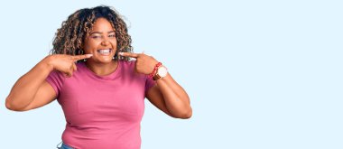 Genç Afro-Amerikan artı cüsseli kadın günlük kıyafetler giyiyor neşeli bir şekilde gülümsüyor ve parmaklarıyla ve ağzıyla işaret ediyor. diş sağlığı kavramı. 