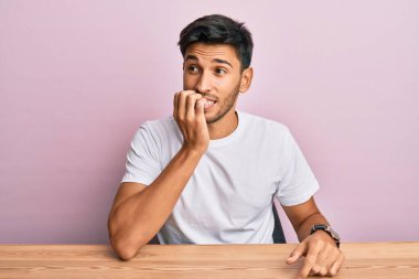 Masada oturan sıradan beyaz tişört giyen yakışıklı genç adam gergin ve gergin görünüyor. Ağzı tırnak yiyen eller. anksiyete. 