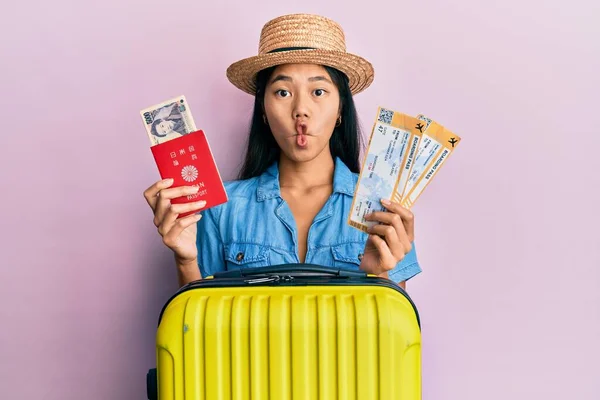 Young chinese woman holding boarding pass and passport making fish face with mouth and squinting eyes, crazy and comical.