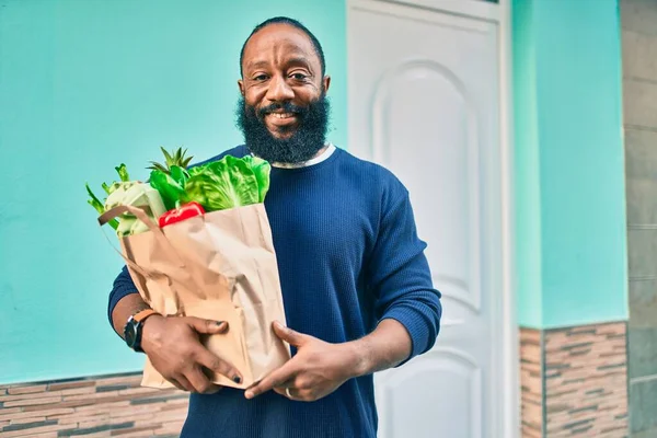 Sakallı Afrikalı Amerikalı bir adam elinde süpermarketten alınmış kağıt poşetler tutuyor.