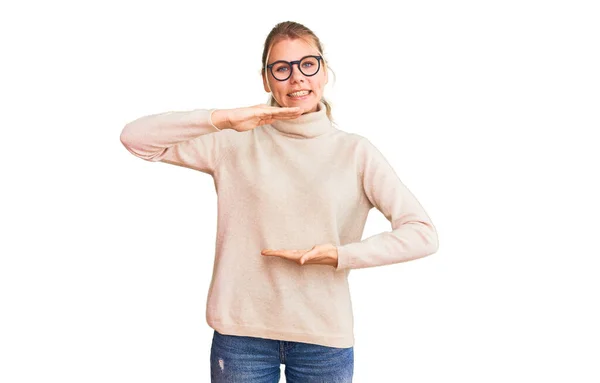 大小のサインを示す手でジェスチャータートルネックセーターや眼鏡を身に着けている若い美しいブロンドの女性は シンボルを測定します カメラを見て笑ってる 概念を測定し — ストック写真