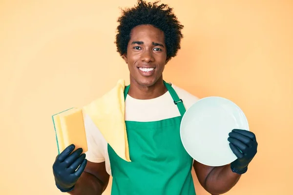 英俊的非洲裔美国男人 头戴围裙 头戴围裙 手里拿着洗碗器 脸上挂着快乐而清凉的笑容 微笑着洗碗 显示牙齿 — 图库照片