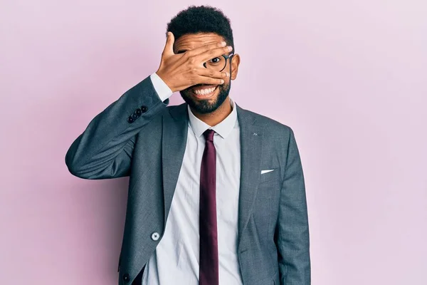 Handsome hispanic business man with beard wearing business suit and tie peeking in shock covering face and eyes with hand, looking through fingers with embarrassed expression.