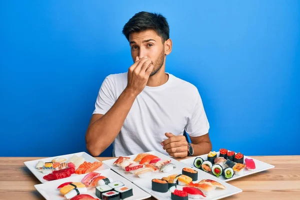 Young handsome man eating sushi sitting on the table smelling something stinky and disgusting, intolerable smell, holding breath with fingers on nose. bad smell