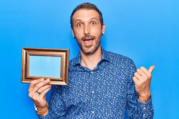 Young Handsome Man Holding Empty Frame Pointing Thumb Side Smiling — Stock Photo, Image