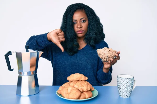 テーブルの上に座っている美しいアフリカ人女性怒り顔のシリアルを食べます 親指で嫌いを示す負の記号を下に示します 拒絶概念 — ストック写真