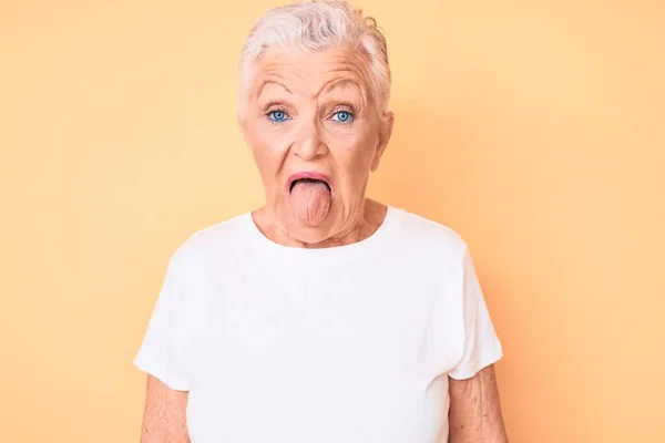 年长美丽的女人 蓝眼睛 灰白的头发 穿着经典的白色T恤 黄背景 带着滑稽的表情伸出舌头 情感概念 — 图库照片