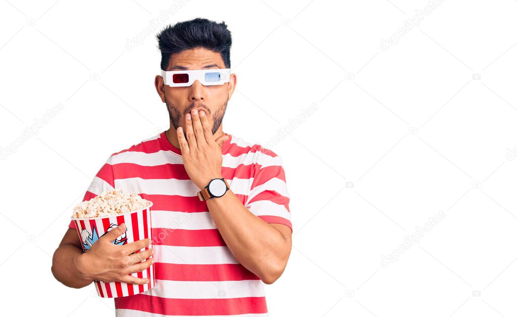 Handsome latin american young man wearing 3d glasses and eating popcorn at the movies covering mouth with hand, shocked and afraid for mistake. surprised expression 