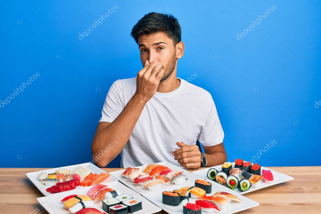 Joven hombre guapo comiendo sushi sentado en la mesa oliendo algo
