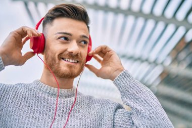 Genç beyaz adam gülümsüyor, şehirde kulaklık takıp müzik dinliyor..