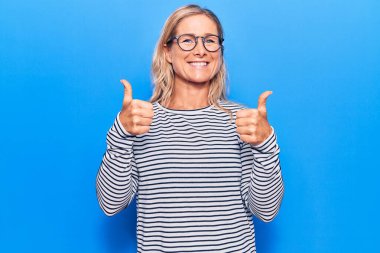 Orta yaşlı, beyaz, çizgili süveter ve gözlük takan sarışın bir kadın. Başarının işareti. Elleriyle olumlu jest yapıyor. Neşeli ifade ve kazanan jest. 