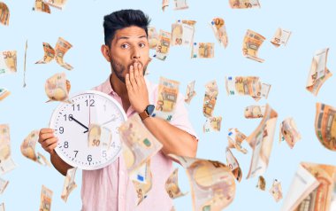 Yakışıklı Latin Amerikalı genç adam büyük bir saat tutuyor. Ağzını eliyle kapatıyor. Şok olmuş ve hata yapmaktan korkuyor. şaşırmış ifade