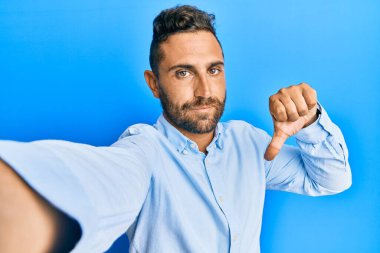 Sakallı yakışıklı bir adam akıllı telefonlu, kızgın yüzlü bir selfie çekiyor. Eksi işareti baş parmakları aşağıda, reddedilme konsepti ise reddedilmiş. 