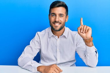 Yakışıklı İspanyol bir adam iş kıyafetleri içinde masaya oturmuş bir numaralı parmağını gösterirken kendinden emin ve mutlu gülümsüyor.. 