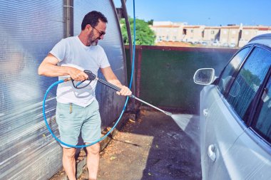 Orta yaşlı yakışıklı, günlük kıyafetler giyen ve mutlu gülümseyen güneş gözlüklü bir adam. Yüzünde gülümsemeyle ayakta durup su tabancası kullanarak araba yıkamak..