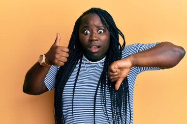 Örgülü genç siyahi kadın baş parmağını kaldırıyor. Korkmuş ve şaşırmış bir şekilde hareket ediyor. İfade, korku ve heyecanlı bir yüz ifadesi var.. 
