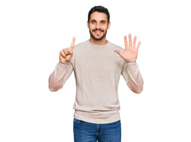 Latin asıllı genç adam gündelik giysiler giyip altı numaralı parmaklarıyla işaret ederken kendinden emin ve mutlu bir şekilde gülümsüyor.. 