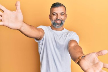 Orta yaşlı, beyaz tişört giyen yakışıklı bir adam kameraya sarılmak için gülümseyerek bakıyor. Mutluluğu kucaklayan neşeli bir ifade.. 