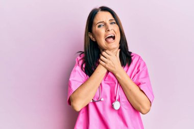 Orta yaşlı esmer kadın doktor üniforması giyiyor ve steteskop bağırıyor ve boğularak ölüyor çünkü acı verici. Sağlık problemi. Boğulma ve intihar konsepti. 