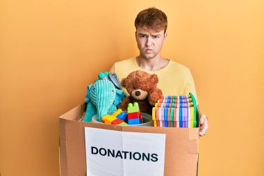 Kafkasyalı genç bir adam oyuncaklarla dolu bir bağış kutusu tutuyor şüpheci ve gergin, sorun yüzünden somurtuyor. negatif kişi. 